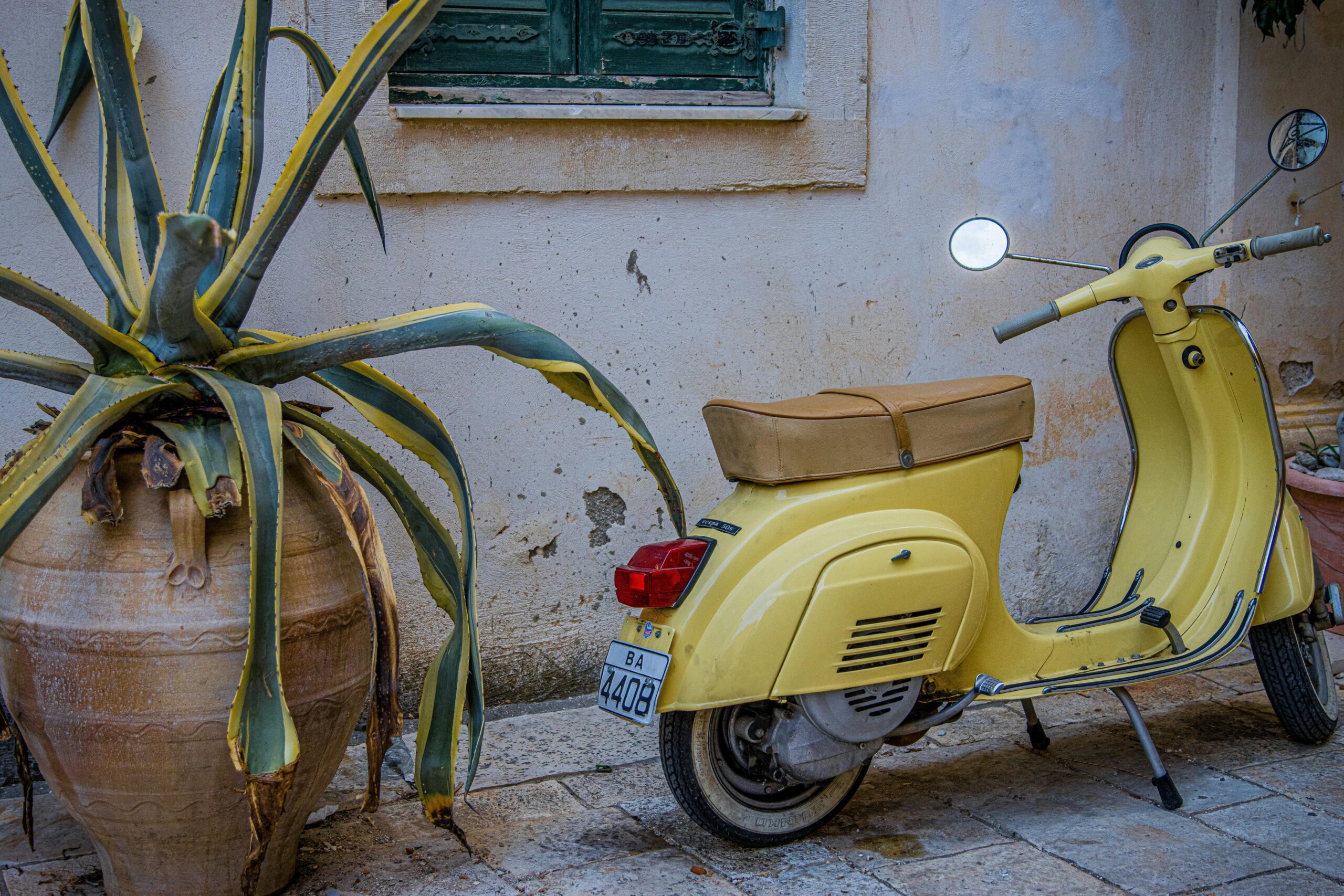 Classic yellow Vespa parked beside potted plant against aged wall in Greece.